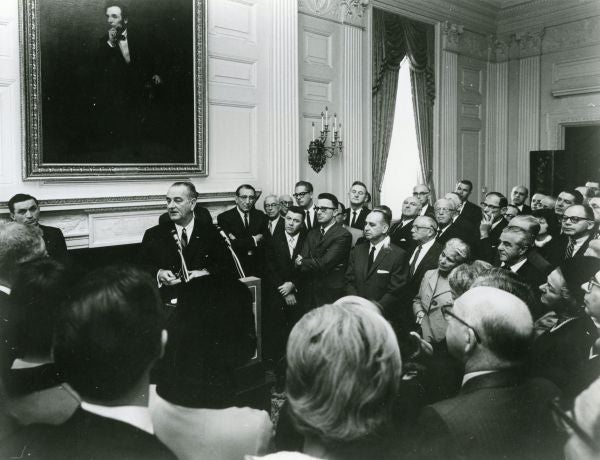 Max M. Fisher looks on as President Lyndon B. Johnson speaks at the White House.